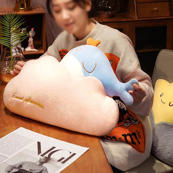 A woman is sitting at a table holding a cloud shaped whale pillow.