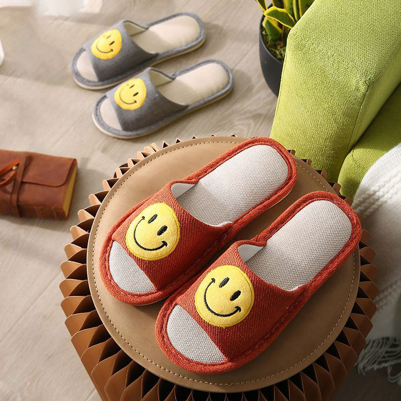 2 pairs of smile face comfortable slippers. The red slipper is on the chair, and the grey slipper is on the floor. On the slipper, there is a yellow smile face on it.